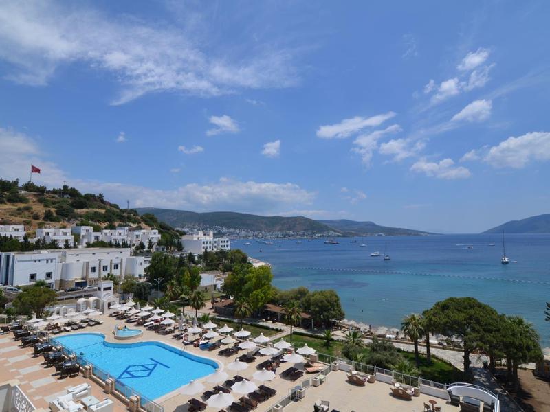 Hotel mit großem Pool, Liegen, weißer Fassade und Blick auf das blaue Meer und den bewölkten Himmel.