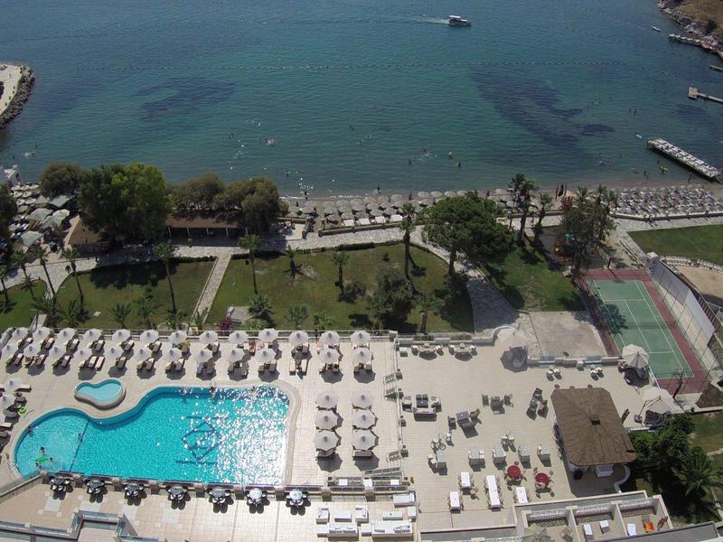 Blick auf ein großes Hotelgelände mit Pool, Liegen, grünen Bäumen und Meer im Hintergrund.