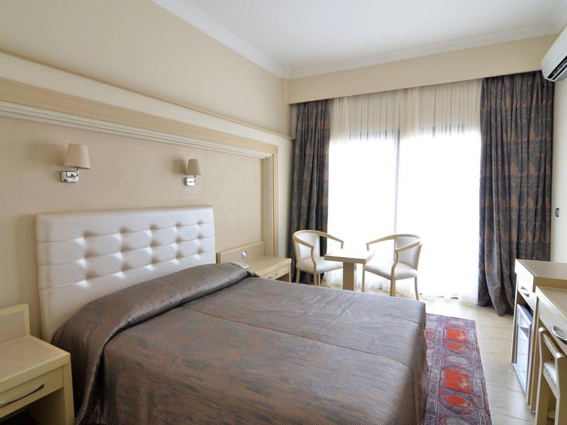 Helles Schlafzimmer mit großem Bett, zwei Stühlen, Schreibtisch, Vorhängen und Klimaanlage an der Wand.