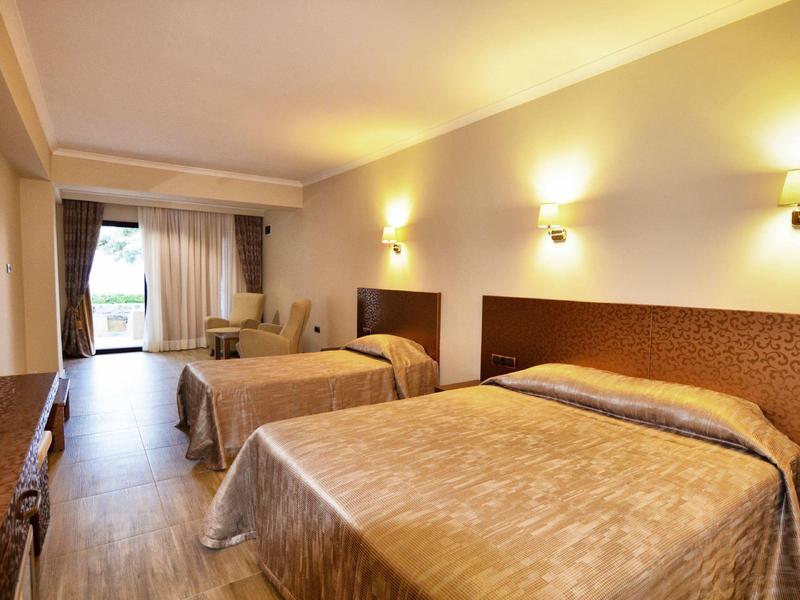 Helles Hotelzimmer mit zwei Betten, Holzboden, Wandlampen und Zugang zu Balkon mit Gartenblick.