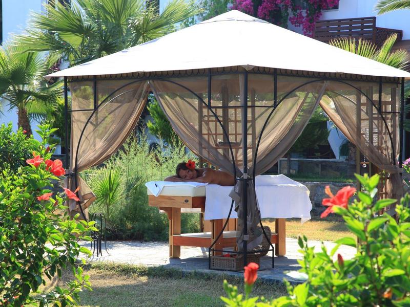 Gartenzelt mit weißem Dach und beigen Textilvorhängen, umgeben von grünen Pflanzen und roten Blüten.