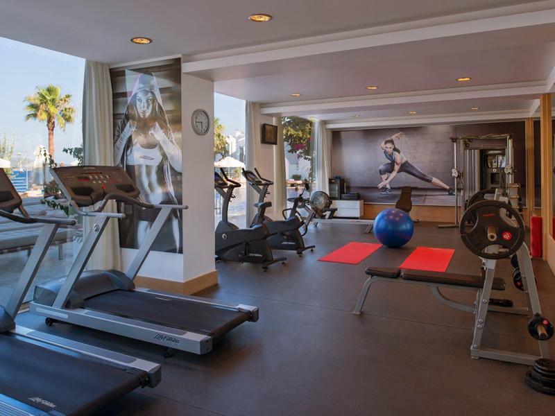 Gimnasio moderno con cintas para correr, pesas y grandes ventanas con vista a palmeras.