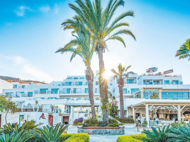 Hotel con edificios blancos y palmeras bajo un cielo azul, rodeado de plantas verdes.