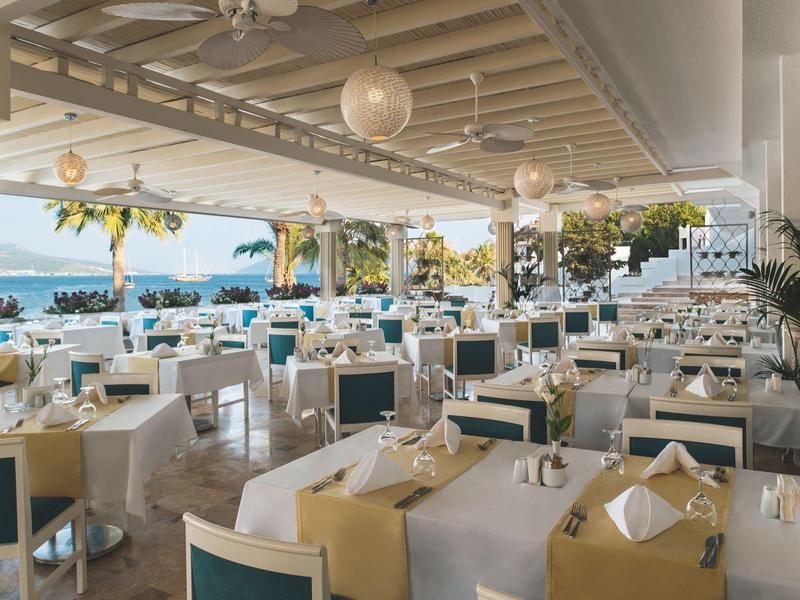 Restaurante abierto de hotel con mesas y sillas blancas, vista al mar y palmeras al fondo.