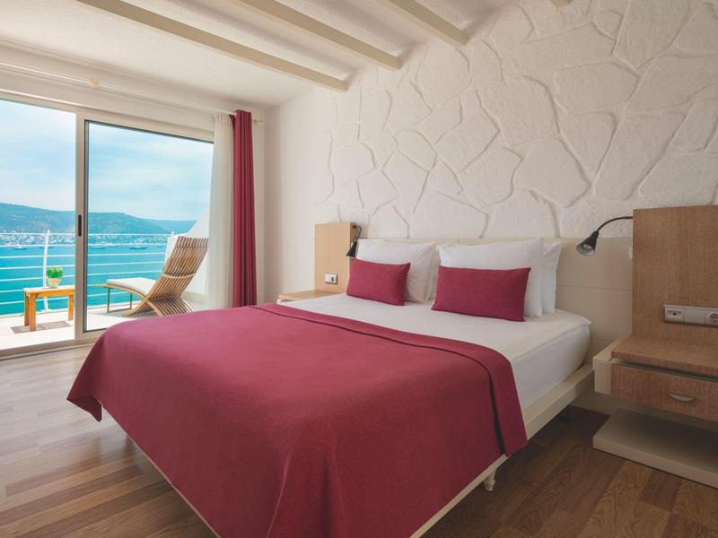 Dormitorio con cama doble, colcha roja, vista al mar y balcón con sillas y mesa.