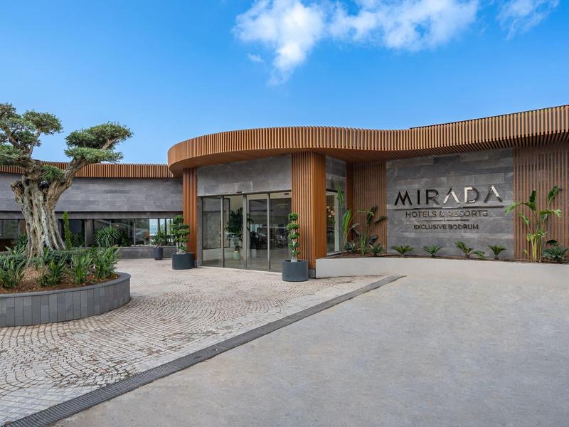 Nowoczesne wejście do hotelu z roślinami i szklaną fasadą pod częściowo pochmurnym niebem.