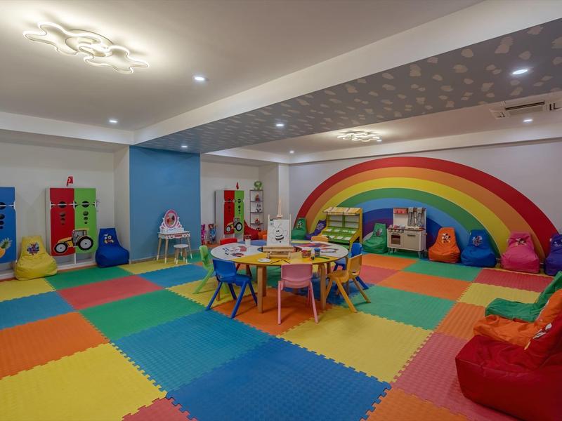 Kolorowa sala zabaw z matami, stolikami, krzesłami i tęczą na ścianie.