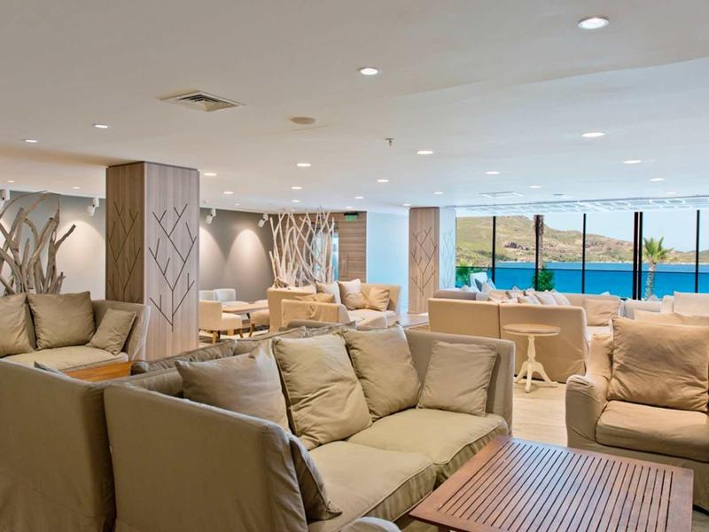 Sala de estar amplia y moderna con sofás beige y vista al mar a través de grandes ventanas.