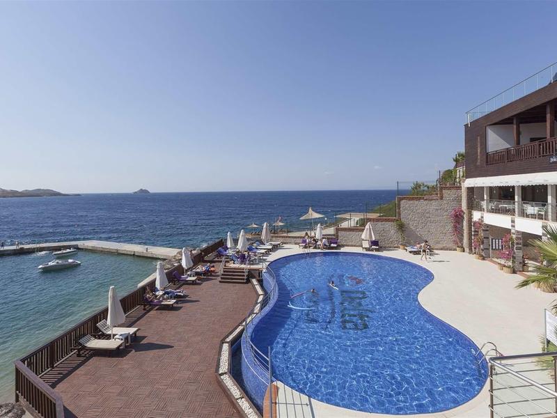 Vista de una piscina con tumbonas junto al mar bajo un cielo azul claro.