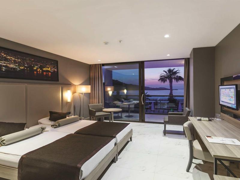 Habitación de hotel moderna con dos camas, escritorio, televisor y vista al mar al atardecer.