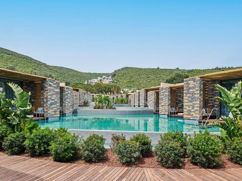 Modern outdoor pool with stone walls and lounge areas surrounded by green hills.