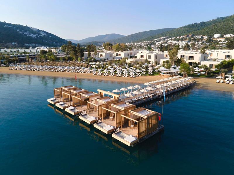 Beach resort with white buildings, sun umbrellas, and wooden pavilions on a pier over blue water.