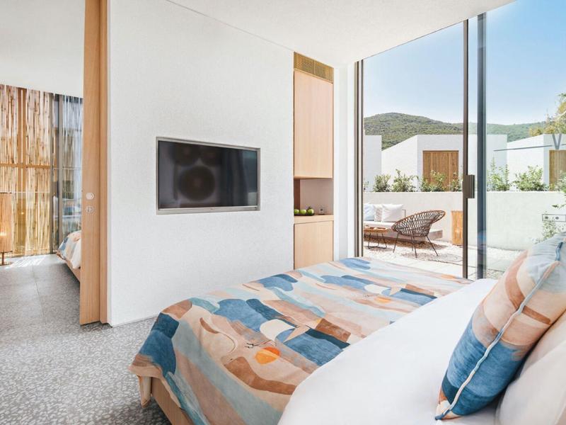 Bright hotel room with bed, wall-mounted TV, and access to terrace overlooking outdoor area.