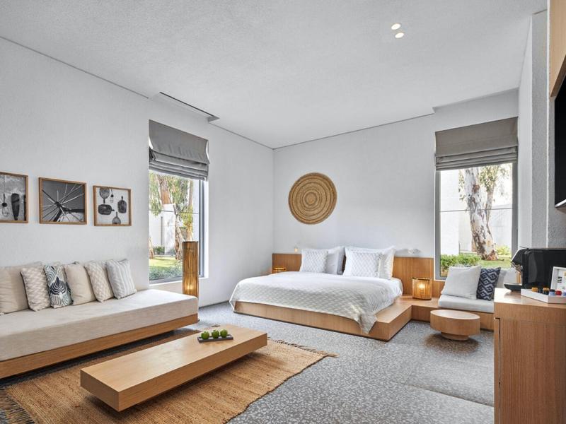 Bright hotel room with double bed, sofa, wooden furniture, and large window overlooking garden.