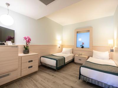 Helles Hotelzimmer mit zwei Einzelbetten, Nachttisch, Kommode und moderner Beleuchtung.
