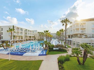 Modern hotel complex with multiple pools, sun loungers, and palm trees under a blue sky.