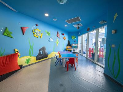 Bunter Spielraum mit Kindern, Tischen und Stühlen, Wandmalerei und großen Fenstern.