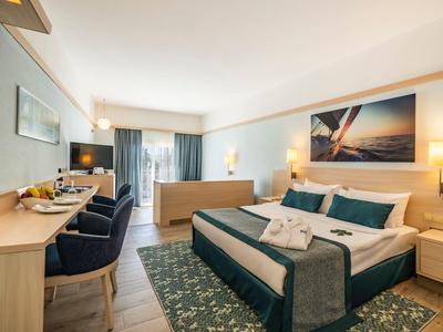 Modern hotel room with large bed, desk, armchairs, and balcony overlooking the sea.