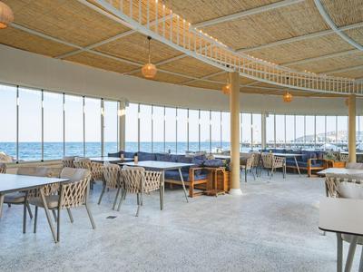 Offenes Restaurant mit vielen Stühlen und Tischen, Blick auf das Meer durch große Fenster.