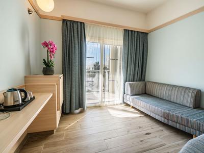 Helles Hotelzimmer mit grauer Couch, Holzmöbeln und Balkon mit Blick ins Freie.
