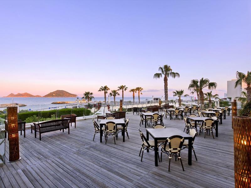 Outdoor terrace with tables and chairs, palm trees, and a sunset over the sea in the background.