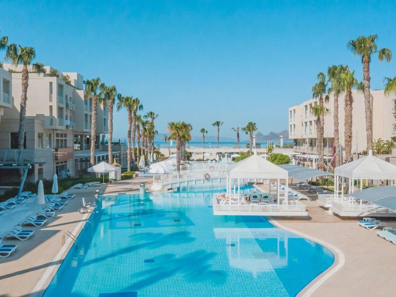 Large outdoor pool with sun loungers and palm trees between hotel buildings under clear sky.