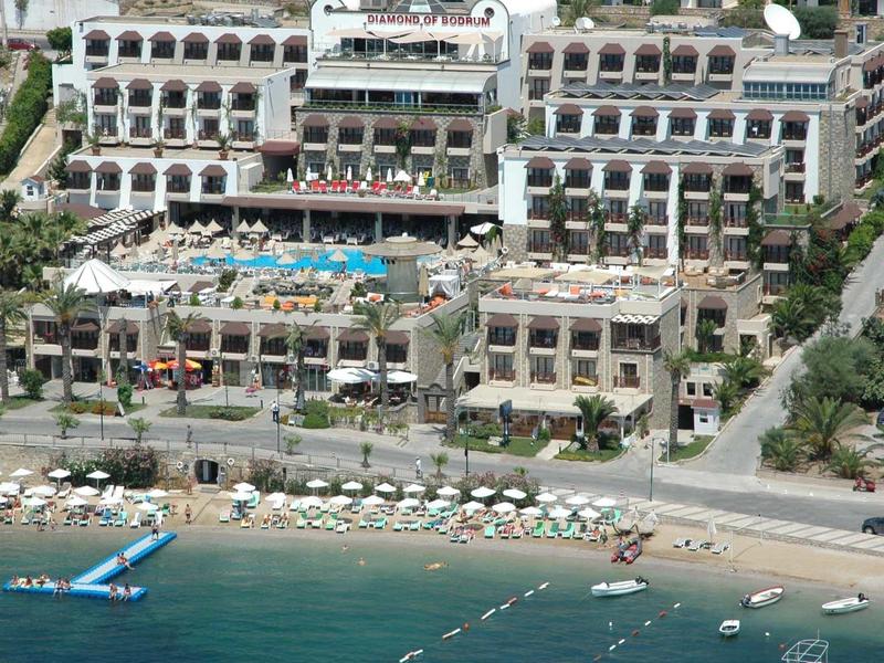 Großes Hotel mit mehreren Stockwerken, Pool, Sonnenliegen und Strand mit klarem, blauem Wasser.