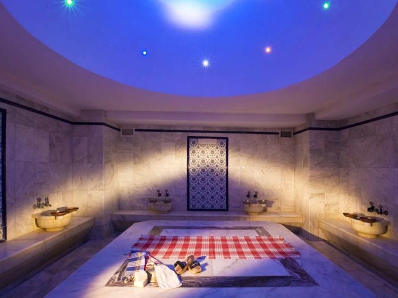 Großer Raum mit Steinwänden, Teppich in der Mitte, rundem blauem Deckenlicht und Wandbrunnen.