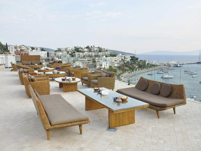 Moderner Lounge-Bereich mit braunen Sofas und Tischen auf Dachterrasse mit Meerblick und Booten.