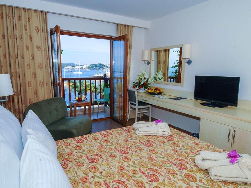 Helles Hotelzimmer mit Doppelbett, dunkelgrünem Sessel, TV, Balkon und Meerblick.