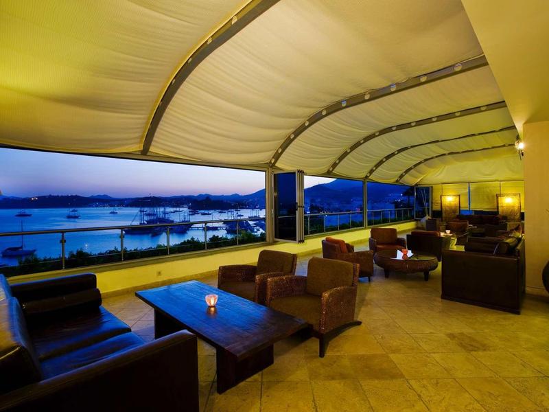 Große Terrasse mit gepolsterten Sesseln, Tischen und Blick auf Wasser bei Abenddämmerung.