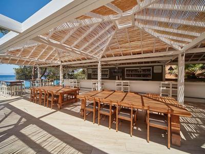 Covered outdoor area with wooden tables and chairs, sea and blue sky in background.