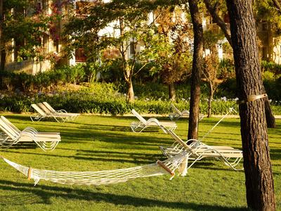 An empty white hammock chair hangs between two trees on a green lawn with lounge chairs.