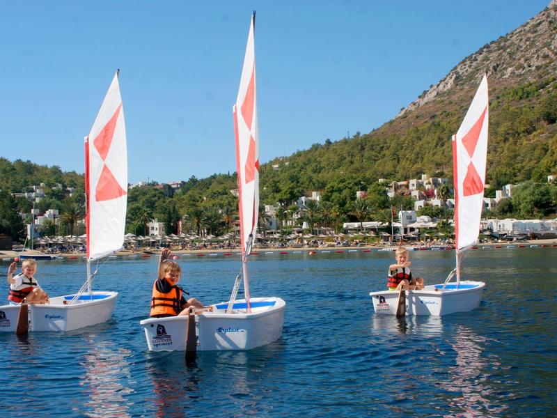 Three people in small sailboats on calm water with wooded hills in the background.