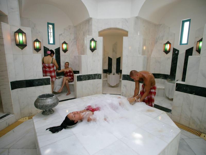 Traditional hammam with marble platform, two people, and glowing wall lanterns.