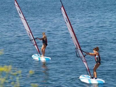 Dos personas haciendo windsurf en pequeñas tablas de vela sobre agua tranquila.
