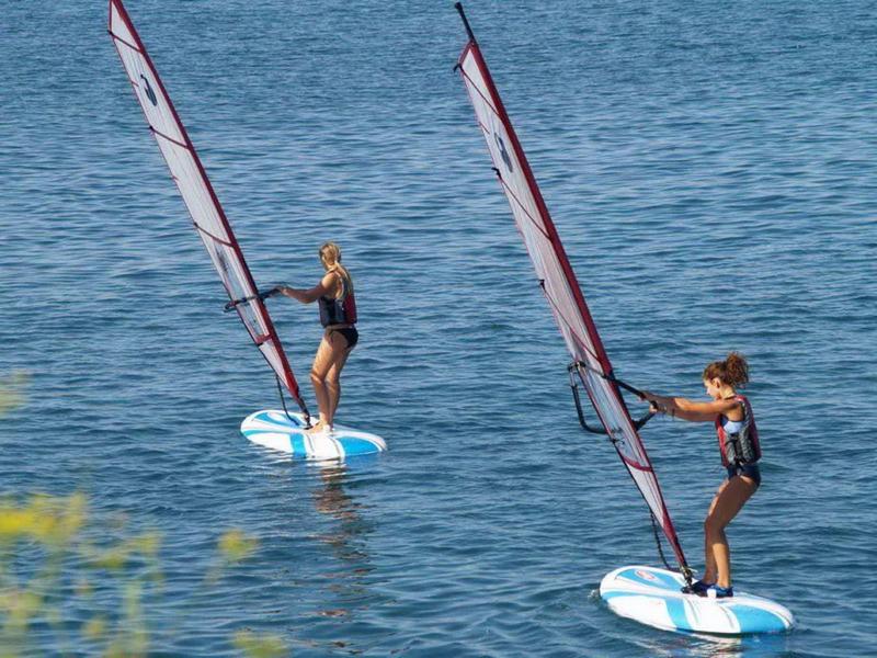 Zwei Personen auf Hydro-Foil-Boards mit Segeln gleiten über ruhiges, blaues Wasser.