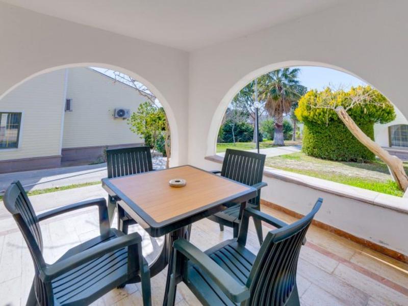 Überdachte Terrasse mit Tisch und vier Stühlen, Blick auf Garten und Häuser mit grüner Bepflanzung.