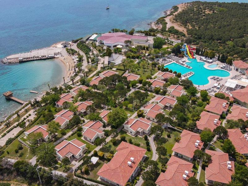 Rote Dächer, großer Pool und Meer in einer grünen Küstenanlage mit Strand und Steg.