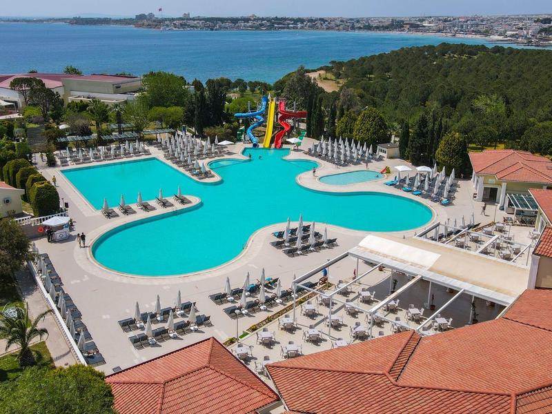 Großer blauer Pool mit Wasserrutschen, Liegestühlen und roten Dächern in grüner Landschaft.