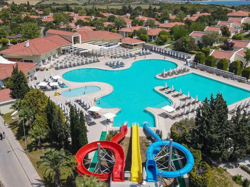 Großer blauer Pool mit Wasserrutschen und Liegestühlen inmitten von grünen Bäumen und Gebäuden.