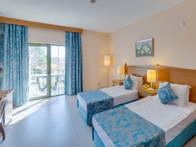 Helles Hotelzimmer mit zwei Betten, blauen Vorhängen und Balkon mit Blick nach draußen.