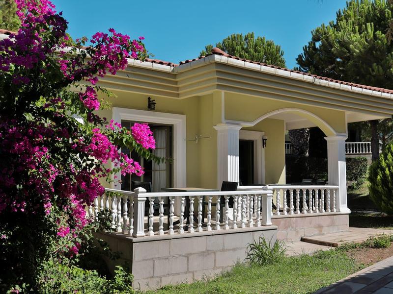 Ein gelbes Haus mit weißem Balkon, blühendem pinken Strauch und grünem Garten bei blauem Himmel.