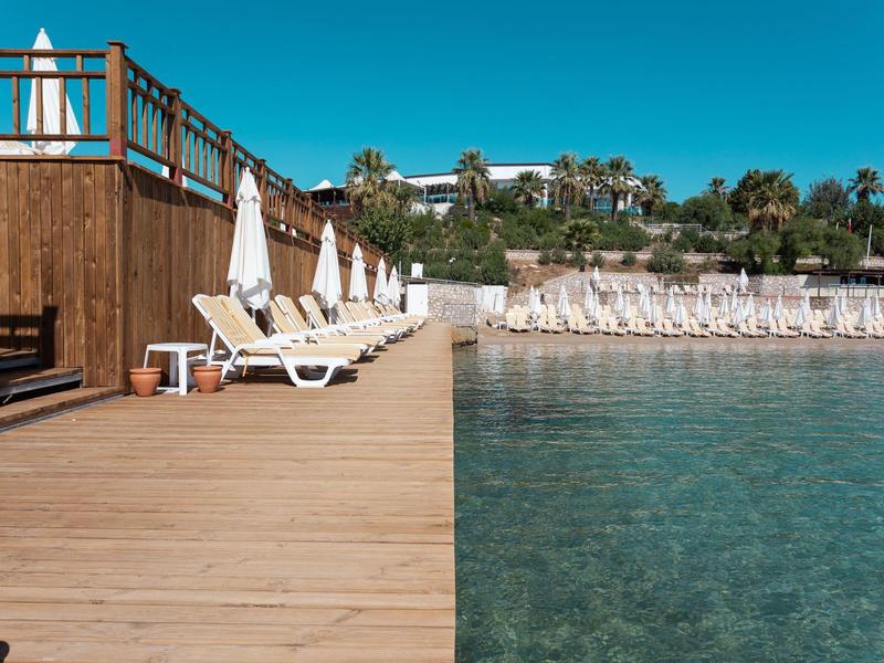 Holzsteg mit Liegestühlen und Sonnenschirmen an klarem Wasser unter blauem Himmel.