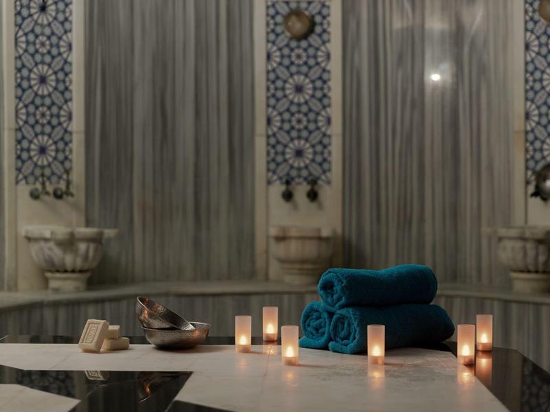 Innenraum eines Hamams mit Marmorwänden, blauen Handtüchern und brennenden Kerzen auf weißer Bank.
