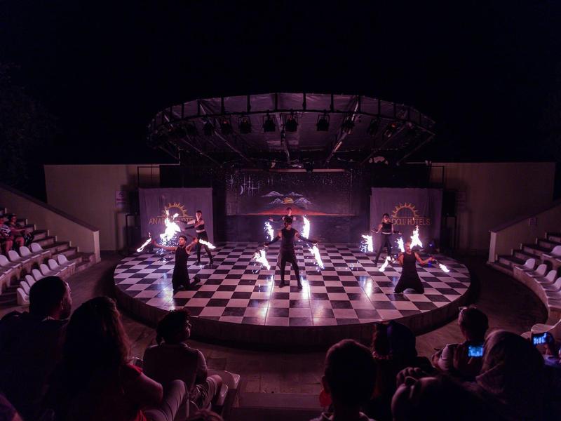 Theaterbühne mit sechs kostümierten Darstellern auf schwarz-weißem Schachbrettmuster, Publikum im Dunkeln.