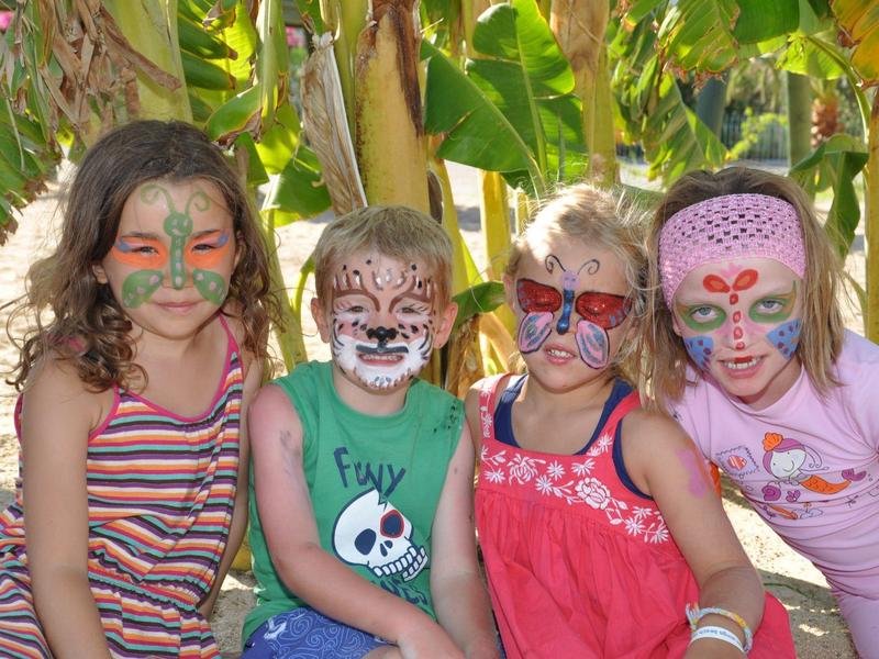 Vier Kinder sitzen vor Bananenpflanzen, alle mit farbenfroher Gesichtsfarbe und Sommerkleidung.