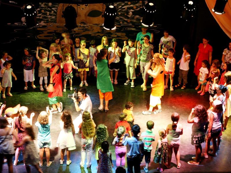 Viele Menschen stehen im Kreis auf einer Bühne, bunte Kleidung, buntes Bühnenlicht.