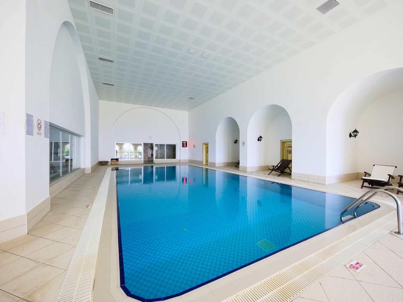 Piscina coperta luminosa con acqua blu chiara, piastrelle beige e pareti bianche con archi.