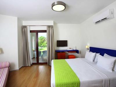 Bright hotel room with double bed, green bed runner, and balcony door overlooking greenery.